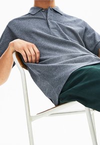 Šedé polo tričko s texturovanou tkaninou, s dvouknoflíkovou légou a krátkými rukávy. V kombinaci s tmavě zelenými kalhotami, sedící na židli.