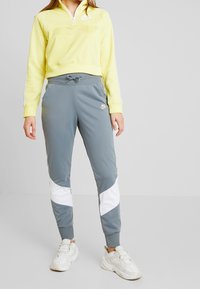 Gul kvartdraget fleece-tröja med hög krage och en vit logotyp. Grå joggingbyxor med vit och grå färgblockering, dragsko i midjan och muddar.