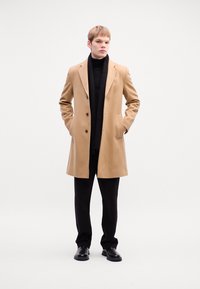 Camel-colored wool overcoat with a tailored design, featuring notched lapels and two front pockets, worn over a black turtleneck and pants.