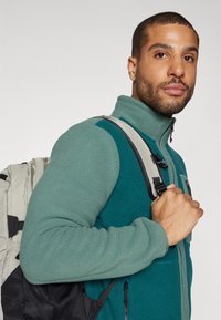 Grønn fleece jakke med glidelås og kontrastfargede teal-detaljer, med en teksturert overflate, samt en grå ryggsekk med svarte detaljer.