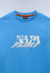 Μπλε βαμβακερό T-shirt με στρογγυλή λαιμόκοψη και πορτοκαλί εσωτερική λεπτομέρεια. Στο εμπρός μέρος εμφανίζεται ένα στυλιζαρισμένο, σκιασμένο λευκό γραφικό "NADA".