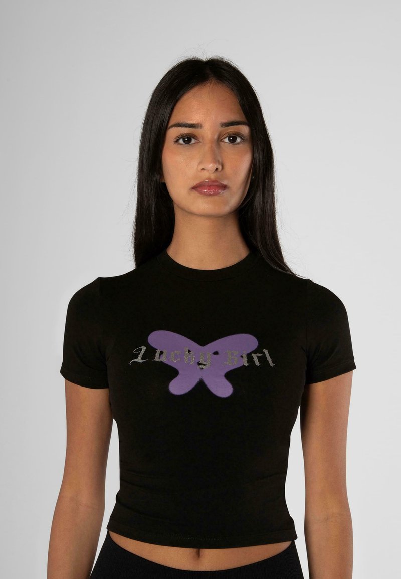 Camiseta negra recortada con un gráfico de mariposa morada y texto "Chica Suerte" en plateado. Mangas cortas con un diseño ajustado.