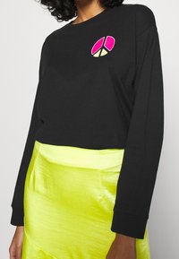 Schwarzes Langarmshirt mit einem pinken und gelben Peace-Symbol-Design auf der Brust, kombiniert mit einem glänzenden neongelben Rock.