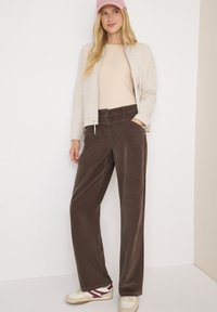 Pantaloni a gamba larga in velluto a coste marroni con una vestibilità comoda, abbinati a una maglietta beige chiaro e a una giacca zip leggera grigia. Sneakers bianche con dettagli rossi.