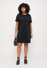 Black short-sleeve dress made of soft material, with a loose fit. The model carries a black bag featuring silver eyelets and wears open-toe black shoes.