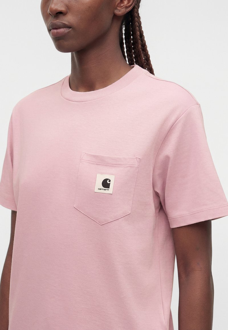 Close-up of a person wearing a light pink short-sleeve t-shirt with a chest pocket featuring a small Carhartt logo patch.