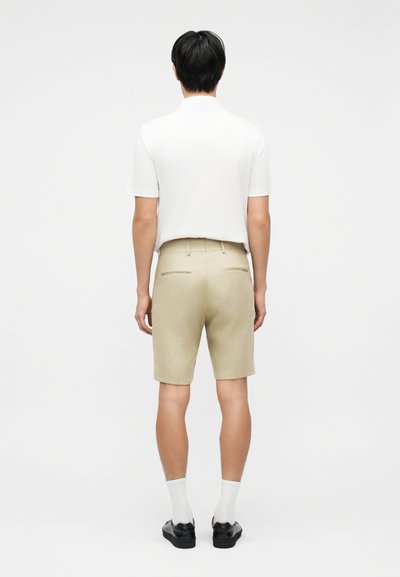 Άνδρας που στέκεται γυρισμένος με την πλάτη, φοράει λευκό κοντομάνικο πουκάμισο, μπεζ σορτς, λευκές κάλτσες και μαύρα παπούτσια σε απλό φόντο.