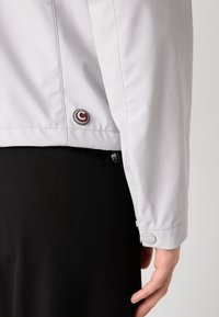 Ανοιχτό γκρι μπουφάν με λεία υφή και εφαρμοστή σχεδίαση. Περιέχει ένα στρογγυλό λογότυπο και μια ασφαλή τσέπη με φερμουάρ στο πλάι.