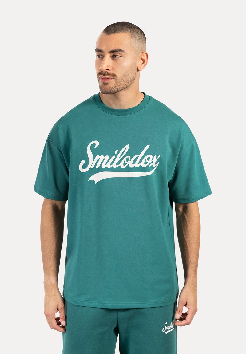 Camiseta de algodón turquesa con mangas cortas, cuello redondo y logo blanco de "Smilodox" en la parte delantera. Textura suave, corte relajado.