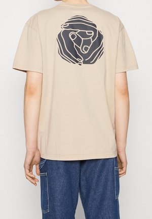 Person trägt beige T-Shirt mit schwarzem abstraktem Hand-Design auf dem Rücken, kombiniert mit blauer Jeanshose mit Seitentaschen.