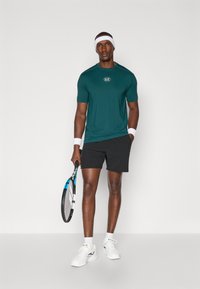 Conjunto atlético masculino com uma t-shirt de manga curta numa tonalidade de teal com um logótipo, calções pretos, sapatos brancos e uma raquete de ténis. Fita branca para a cabeça e fitas de pulso.