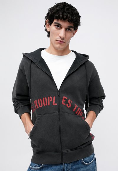 Hanoracul negru cu fermoar, având text roșu imprimat pe piept, dotat cu o buzunar frontal și șnururi. Purtați peste un tricou alb.