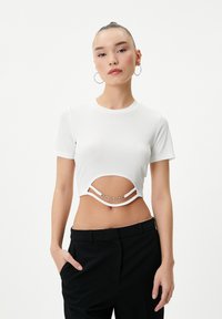 Hvid ribbet crop top med korte ærmer, der har et udskæringsdesign og sølvkædeudsmykninger under brystet. Sammen med sorte bukser.