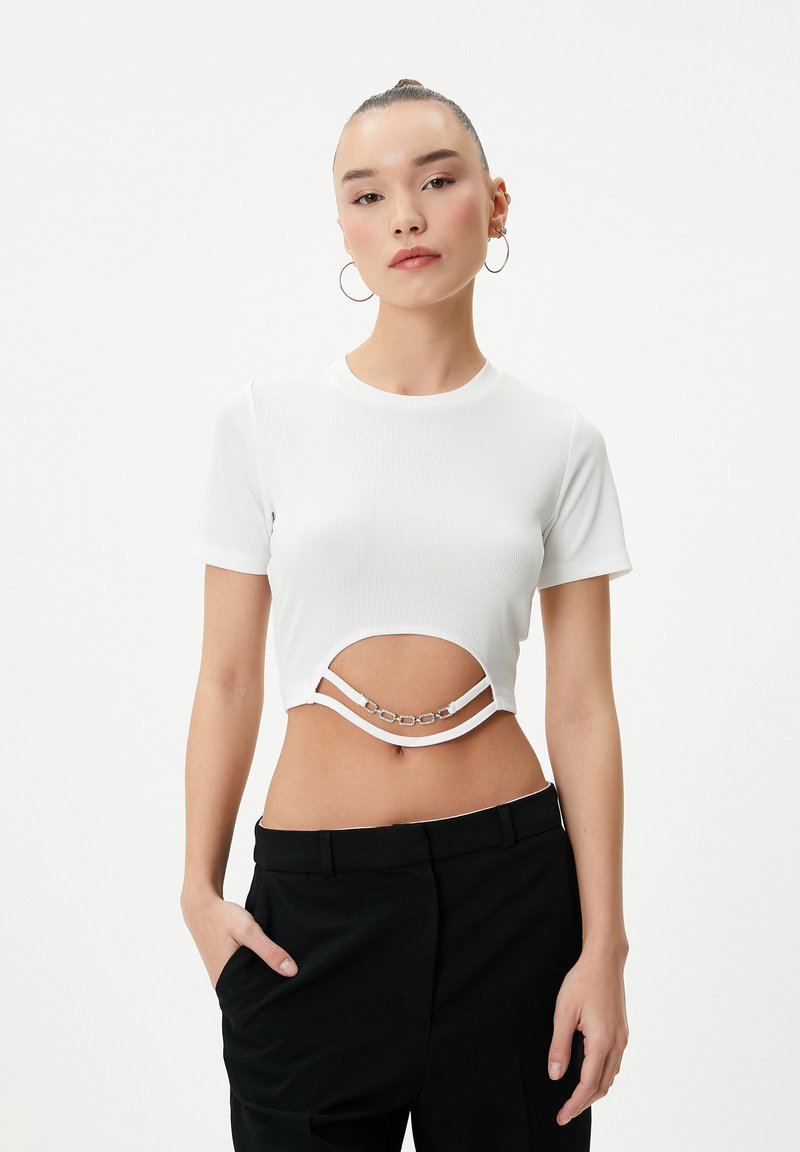 Hvid ribbet crop top med korte ærmer, der har et udskæringsdesign og sølvkædeudsmykninger under brystet. Sammen med sorte bukser.