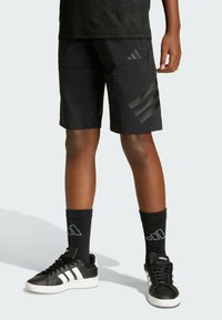 Osoba nosi crne Adidas šortse sa tri pruge, crne Adidas čarape i crne Adidas patike s bijelim đonom.