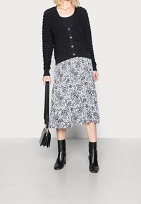 Woman wearing black textured cardigan, black top, white and navy patterned midi skirt, black ankle boots, holding black handbag by side.