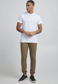 Camiseta polo blanca con tapeta de tres botones, combinada con pantalones jogger color caqui y zapatillas negras, en un fondo gris claro.