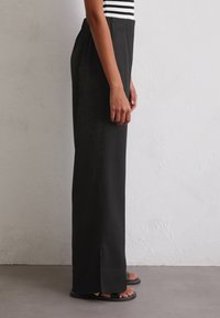 Pantalons larges noirs avec une taille élastique, texture lisse et fentes sur les côtés ; associés à des sandales noires sur un sol gris.