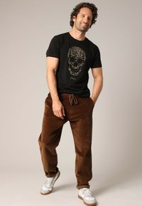 T-shirt en coton noir avec un graphique de crâne doré, associé à un pantalon en velours côtelé marron et des baskets blanches avec des accents marron.