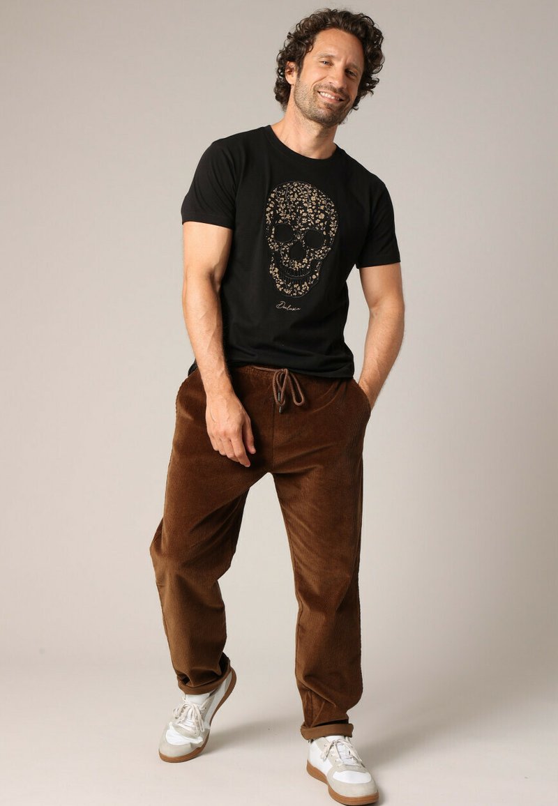 T-shirt en coton noir avec un graphique de crâne doré, associé à un pantalon en velours côtelé marron et des baskets blanches avec des accents marron.