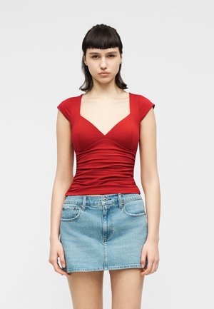 Jauna moteris dėvi aptemptą raudoną viršutinę dalį su gilia iškirpte ir šviesiai mėlyną džinsinę mini sijoną, stovinčią priešais paprastą šviesią fono sieną.