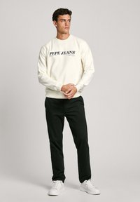 Krémová mikina s černým logem "PEPE JEANS", žebrovanými manžetami a spodním lemem, spárovaná s černými kalhotami a bílými teniskami. Hladká textura látky.