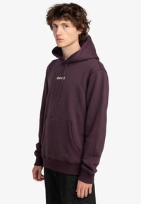 Sudadera morada de mezcla de algodón, con un bolsillo frontal y capucha con cordón. Pequeño logo dorado en el pecho. Textura suave, ajuste casual.