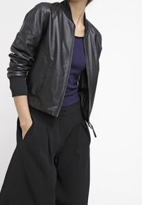 Schwarze Lederbomberjacke mit Rippbündchen und Kragen, ausgestattet mit einem Reißverschluss. Kombiniert mit weiten schwarzen Hosen und einem lila Oberteil.