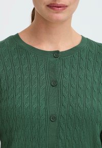 Cardigan en laine verte tricotée avec un motif de câbles texturé, un col rond et deux boutons ronds verts à l'avant.
