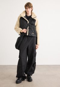 Μαύρο επενδυμένο μπουφάν με καφέ γούνα στο γιακά, φορεμένο πάνω από ένα μαύρο πουκάμισο. Σε συνδυασμό με loose μαύρες cargo παντελόνες και χοντρά μαύρα μποτάκια.