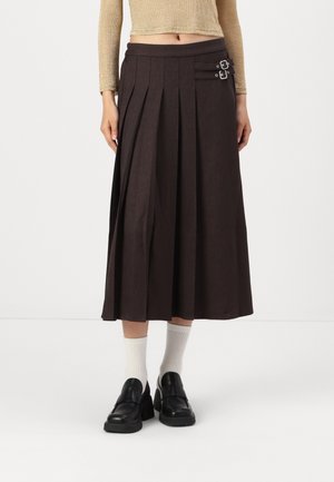 Femme portant une jupe midi plissée marron foncé avec deux boucles latérales, des chaussettes blanches, des mocassins noirs et un top court à manches longues beige.