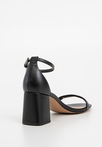 Sandale en cuir noir avec talon bloc, bride à la cheville avec boucle et fine bride avant sur fond blanc uni.