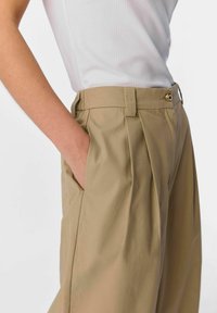 Pantalon kaki à coupe ajustée, avec des plis avant, des poches latérales, une ceinture assortie et des détails en métal doré.