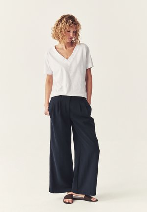 Kobieta o kręconych włosach, ubrana w biały, krótki top z dekoltem w serek, czarne szerokie spodnie i czarne klapki, stoi z rękami w kieszeniach.