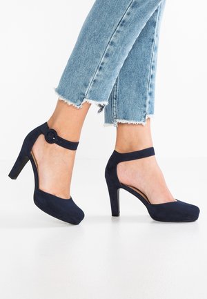 Mørkeblå højhælede sko med en ruskindslignende tekstur, lukket tå og ankelrem med en cirkulær spænde. Sammen med jeans med trådtede kanter.