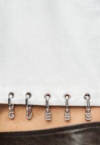 Λευκή βαμβακερή μπλούζα με ασημένιες τρούκς και γράμματα διακοσμημένα με στρας που σχηματίζουν τη λέξη "GUESS" κατά μήκος του τελειώματος, συνδυασμένη με σκούρο υφασμάτινο ύφασμα με υφή.