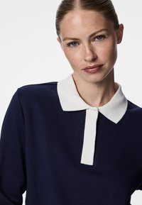 Mørkeblå langermet polo-skjorte med hvit krage og knappestolpe, laget av et glatt stoff med en tettsittende design og minimalistiske detaljer.