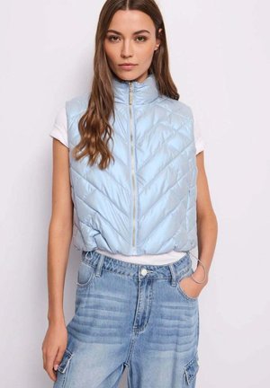 Donna con gilet trapuntato senza maniche azzurro chiaro sopra una t-shirt bianca e jeans cargo blu a vita alta con la mano in tasca.