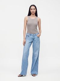 Femme debout portant un haut gris sans manches côtelé, un jean large bleu clair et des sandales noires ouvertes à bout ouvert sur un fond clair uni.