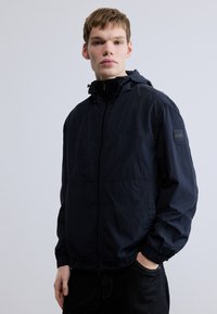 Tmavomodrá bunda na zips s kapucňou, elastickými manžetami a jemným leskom. Na ľavom rukáve má logo nášivku. Ľahká, hladká textúra.