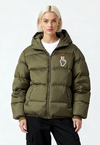Veste matelassée vert olive avec capuche et fermeture éclair, arborant un graphisme de main blanche avec un cœur rouge sur la poitrine. Tissu texturé et matelassé.