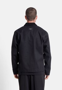 Giacca nera in tela, con collo e un'etichetta sul retro, caratterizzata da cuciture in vista. Tessuto liscio, design minimale.