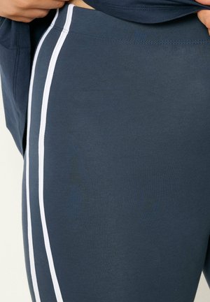 Close-up van marineblauwe leggings met twee verticale witte strepen aan de zijkant, gedragen door een persoon die hun tailleband aanpast.