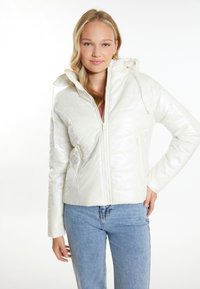 Giacca puffa bianca, trapuntata, con finitura lucida, cappuccio, chiusura frontale con zip e tasche laterali. Indossata con jeans azzurri chiari.
