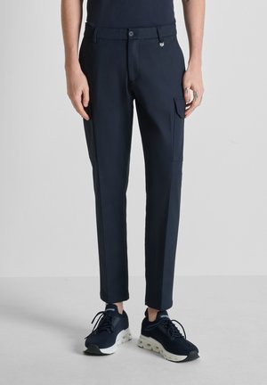 Hombre con pantalones negros de corte ajustado con bolsillo lateral y zapatillas negras con suelas blancas, de pie sobre un suelo gris.