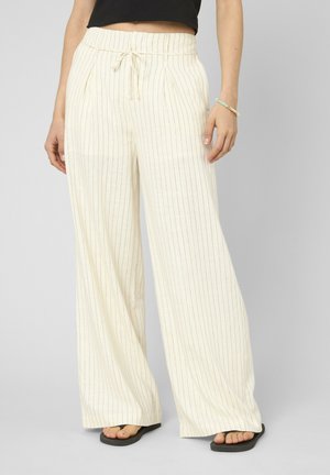 Femme portant un pantalon crème à jambes larges avec de fines rayures noires verticales, un crop top noir, un bracelet et des tongs noires, debout sur un sol gris.