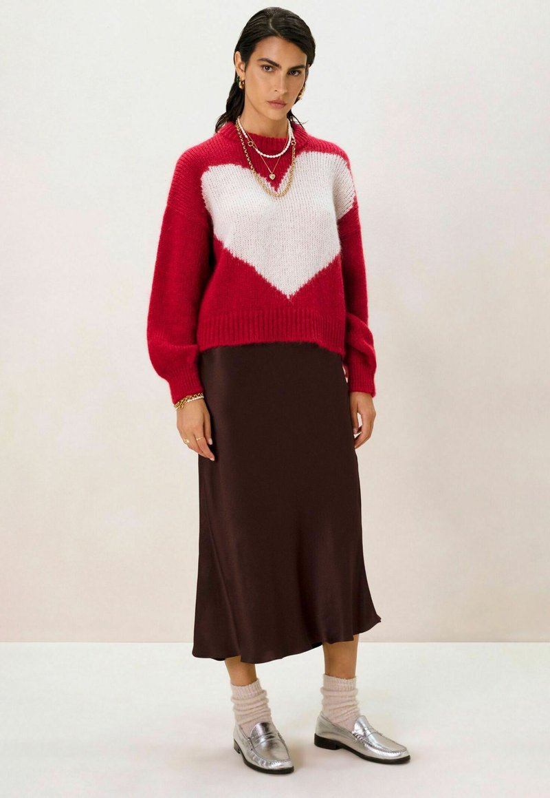 Pull en tricot rouge et blanc avec un design de cœur, jupette satinée marron, chaussures argentées et chaussettes en tricot crème clair. Accents de bijoux simples.