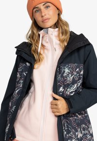 Frau mit einer orangefarbenen Strickmütze, einem rosa Reißverschluss-Fleece und einer schwarzen Jacke mit bunten Mustern, die die Jacke mit einer Hand offen hält.