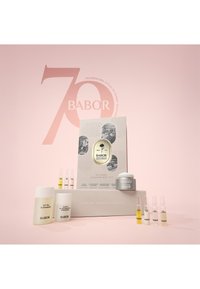 El kit del 70º aniversario de Babor incluye un limpiador blanco, una crema hidratante, ampollas variadas y un frasco gris, presentados sobre un fondo rosa.