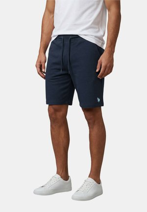 Mann trägt marineblaue Shorts mit Kordelzug, weißes T-Shirt und weiße Turnschuhe, steht vor einem schlichten hellen Hintergrund.